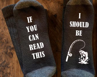 May include: A pair of dark grey socks with white text and graphics. The left sock reads IF YOU CAN READ THIS and the right sock reads I SHOULD BE with a graphic of a fishing rod and a fish jumping out of the water below the text.