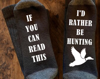 May include: A pair of dark grey socks with white text and a white duck graphic. The left sock features the text "IF YOU CAN READ THIS" and the right sock features the text "I'D RATHER BE HUNTING" above a silhouette of a flying duck.