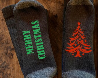 May include: A pair of dark grey socks with a textured knit. One sock features the text MERRY CHRISTMAS in bright green lettering, while the other sock displays a stylised red Christmas tree graphic with star and circular ornaments.
