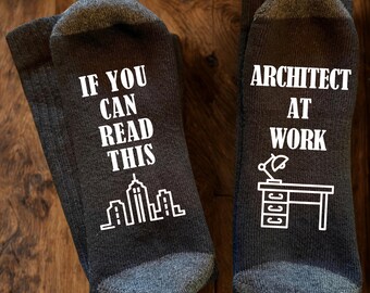 May include: A pair of dark charcoal grey socks with white text and graphics. The left sock reads IF YOU CAN READ THIS above a city skyline. The right sock reads ARCHITECT AT WORK above a graphic of a desk with a lamp and drawers.