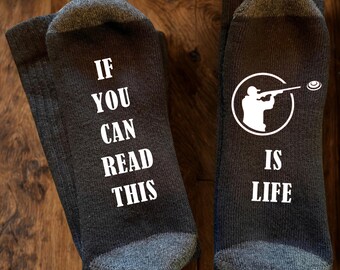 May include: A pair of dark grey socks with white text and graphics. The left sock reads IF YOU CAN READ THIS in white capital letters. The right sock features a white circular graphic of a person shooting a clay target and the text IS LIFE.