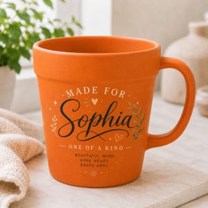 Può includere: Una tazza arancione in stile terracotta con manico, che riporta la scritta "MADE FOR Sophia ONE OF A KIND" in nero. Sotto il nome, il testo recita "BEAUTIFUL MIND KIND HEART BRAVE SOUL" con piccoli decori a forma di foglie, cuori e stelle.