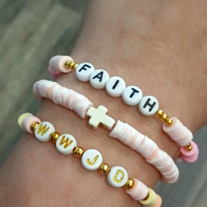 May include: Three handmade bracelets with white letter beads spelling "FAITH" and "WWJD". The bracelets are made with pink and white disc beads, gold accents, and a white cross charm. A stylish accessory.