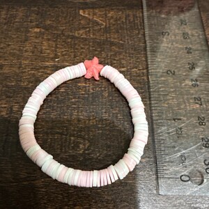 May include: A pastel-coloured bracelet with pink, white, and light green disc-shaped beads. A small, coral-coloured starfish charm is attached. The bracelet is approximately 18 cm in circumference.