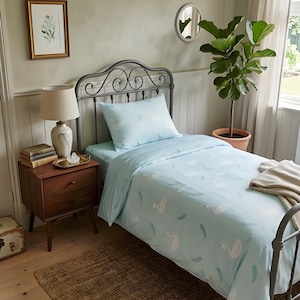 May include: A bedroom scene featuring a bed with a light blue swan-patterned duvet cover and pillowcase. The bed has a wrought iron headboard. A wooden nightstand with a lamp, books, and a decorative tray sits beside the bed. A woven rug is on the floor.