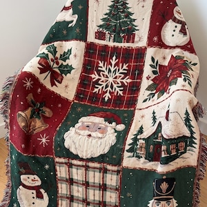 Peut inclure: Un plaid tissé présentant un motif patchwork de thèmes de Noël, incluant un sapin décoré, un bonhomme de neige, un flocon, du houx, des cloches, le Père Noël, un chalet d'hiver et un casse-noisette dans des tons rouge, vert, blanc et brun.