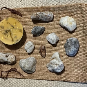 May include: A collection of natural stones and a round, yellow item with green specks, possibly soap or a candle, arranged on a burlap sack. The stones vary in size and color, ranging from gray to white and brown.