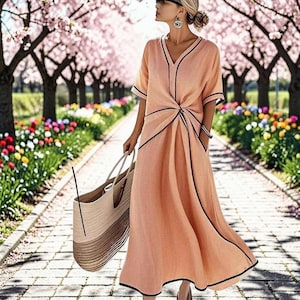 Può includere: Un abito color pesca con rifiniture nere e scollo a V. L'abito ha maniche corte e un dettaglio a nodo in vita. Una borsa tote in paglia con un design a righe è tenuta in mano. Lo sfondo presenta un sentiero fiancheggiato da alberi e fiori.