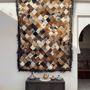 Peut inclure: Un grand tapis rectangulaire composé de carrés cousus de peaux d'animaux dans des tons de marron, beige, noir et blanc. Le tapis a une bordure sombre et hirsute et est accroché à un mur blanc. Un coffre en bois foncé avec des sculptures décoratives est en dessous.