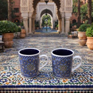 May include: Two ceramic mugs with white handles and rims, featuring a blue and white floral pattern. The mugs are set on a mosaic tile surface, with a blurred background of an outdoor courtyard with arches and palm trees.