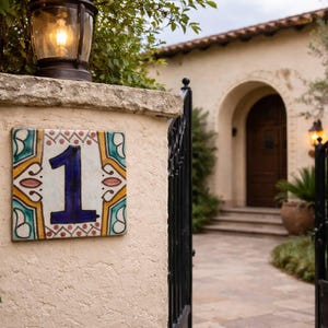 Peut inclure: Une plaque de numéro de maison en céramique décorative avec le chiffre 1 en bleu marine. La tuile a une bordure colorée avec des accents jaunes, turquoise et rouges. Un portail en fer forgé noir et une maison avec une porte en bois sont en arrière-plan.