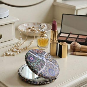 Peut inclure: Une collection de produits de beauté sur une coiffeuse blanche. Comprend un miroir compact à motifs, un rouge à lèvres, un parfum, une palette de fards à paupières, des pinceaux de maquillage et un collier de perles. Les articles sont disposés sur une surface crème.