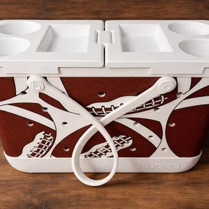May include: A white and brown cooler with a football design. The cooler has a white lid with cup holders and a handle. The cooler is designed to keep items cold and is ideal for picnics and outdoor events.