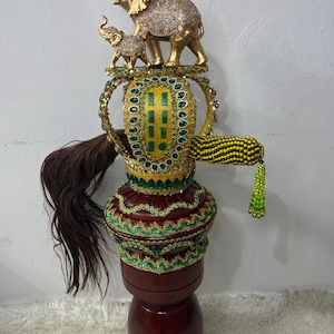 May include: A decorative, ornate sculpture featuring a gold elephant family atop a detailed, jewel-encrusted structure. The sculpture has a dark red base and is adorned with green and gold accents, and a long brown hair-like element.