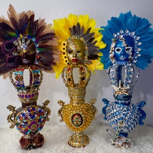 Könnte beinhalten: Drei verzierte Masken mit Kronen und Federhauben. Die Masken sind in Lila, Gelb und Blau gehalten und mit Juwelen und aufwendigen Details verziert. Die Sockel sind mit Edelsteinen und floralen Akzenten verziert.