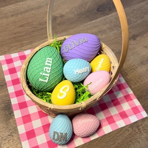May include: A wicker basket containing several brightly coloured, textured Easter eggs. The eggs are in shades of green, purple, blue, yellow, and pink, each with a name or initial. The basket rests on a pink and white gingham cloth.