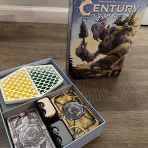 May include: A board game box titled "Century Golem Edition" with a blue and purple illustration of a golem holding a tree. The box is open, revealing game components including cards, tokens, and a two-toned container with yellow and green sections.