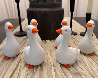 May include: A set of six small white goose figurines with textured bodies, orange beaks, and orange feet. Each goose is depicted in a standing position with a slightly curved neck, featuring black dot eyes and detailed wing feather markings.