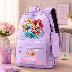 K&ouml;nnte beinhalten: Ein hellvioletter Rucksack mit einer Grafik von drei Prinzessinnen, einer goldenen Krone und dem Namen AIZA auf einem rosa Banner. Die Tasche hat ein Schuppenmuster, eine vordere Rei&szlig;verschlusstasche und ein passendes Etui an der Seite.