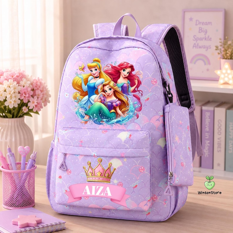 K&ouml;nnte beinhalten: Ein hellvioletter Rucksack mit einer Grafik von drei Prinzessinnen, einer goldenen Krone und dem Namen AIZA auf einem rosa Banner. Die Tasche hat ein Schuppenmuster, eine vordere Rei&szlig;verschlusstasche und ein passendes Etui an der Seite.