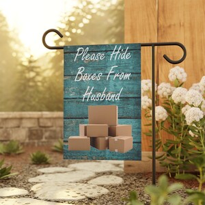 May include: A garden flag with the text "Please Hide Boxes From Husband" on a teal wooden background. The flag features a stack of brown cardboard boxes. The flag is displayed on a black metal stand in a garden setting.
