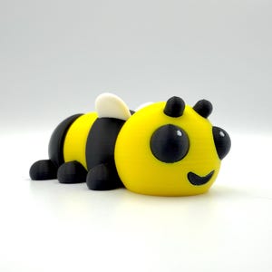 May include: A 3D-printed bee figurine in yellow and black, with a smiling face and small black antennae. The bee has white wings and black feet, and is lying down on a white surface.