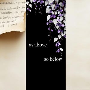 Peut inclure: Marque-page noir avec le texte "as above so below". Le haut du marque-page présente un motif floral avec des fleurs violettes et des feuilles argentées. Le bas du marque-page porte le texte "etherrootspiritualo williamstephaniesawyer".