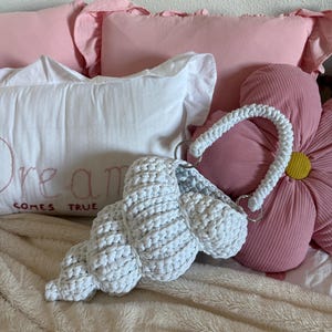 Puede incluir: Un bolso blanco, tejido a ganchillo en forma de concha marina, con un asa blanca y aros plateados. El bolso se encuentra sobre un fondo de almohadas rosas y una almohada blanca con las palabras "Dream Comes True" bordadas. El tema general es una estética de playa o costera.