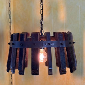 Könnte beinhalten: Eine rustikale, handgefertigte Pendelleuchte. Die Lampe hat dunkelbraune Holzlatten, die von einem Metallband zusammengehalten werden. Eine einzelne Edison-Glühbirne hängt in der Mitte und beleuchtet die Leuchte. Die Leuchte ist an einer schwarzen Kette aufgehängt.
