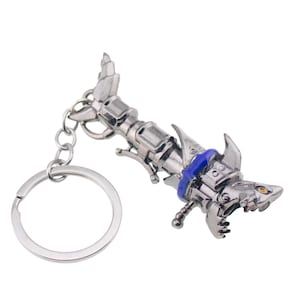 May include: A silver-colored metal keychain featuring a stylized shark design. The shark has sharp teeth, a blue band, and a detailed mechanical appearance. The keychain includes a silver ring for attaching keys.