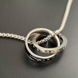 May include: A silver-toned necklace featuring two interlocking rings. One ring is plain silver with the text ETERNAL LOVE engraved on it. The second ring is black with the word Star and small star symbols engraved in white on the outer surface.