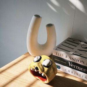 May include: A yellow ceramic face with black and white eyes and an open red mouth sits on a wooden surface. A cream-colored vase with two openings and a stack of books are behind the face.