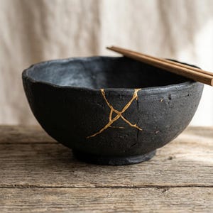 Puede incluir: Un cuenco de cerámica negra con líneas doradas de reparación kintsugi. Dos palillos de madera descansan sobre el borde. El cuenco se encuentra sobre una superficie de madera envejecida, lo que sugiere una estética rústica. El diseño del cuenco es un testimonio del arte japonés de abrazar las imperfecciones.