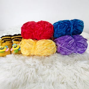 May include: A collection of colorful yarn skeins in red, yellow, blue, and purple. The yarn is soft and plush, perfect for knitting or crocheting projects. Also, there are several small, yellow and black knitted baby booties with smiling bee appliqués.