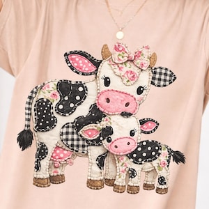 May include: A light pink t-shirt featuring a whimsical design of two cartoon cows. The cows have black and white patterns, pink accents, and floral details. The design is centered on the shirt.