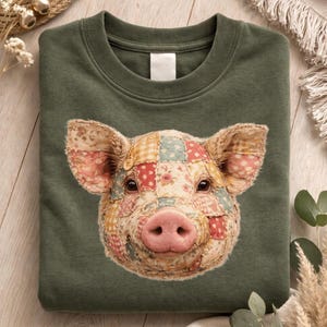 May include: A dark green sweatshirt featuring a patchwork pig face design. The pig's face is made of various fabric patterns in shades of red, pink, yellow, and blue. The pig has a pink nose and brown eyes. The sweatshirt has a crew neck.