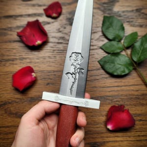 May include: Handcrafted Rose Dagger with a custom engraved blade. The dagger features a silver blade with a rose engraving, a red wooden handle, and a silver guard. The image includes rose petals and leaves.