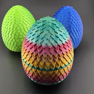 Puede incluir: Tres huevos de dragón decorativos en colores vibrantes. Los huevos son verdes, azules y un huevo multicolor con tonos de rosa, amarillo y verde. Cada huevo tiene un patrón texturizado en forma de escamas. Sobre fondo oscuro.