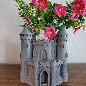 May include: A gray castle-shaped planter featuring two tall side towers, a central arched doorway, and detailed brickwork textures. The structure is filled with artificial green foliage and vibrant pink flowers extending from the top opening.