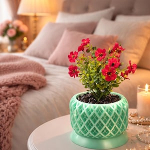 May include: A small, round, mint-green planter with a diamond pattern holds a plant with vibrant red flowers. The planter sits on a white table next to a lit candle and a string of pearls. A bed with pink pillows and a blanket is in the background.