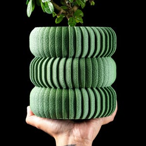 May include: A cylindrical green planter featuring three stacked, ribbed, and textured sections. The exterior surface has a fine, matte, and grooved finish. A small green plant with dark leaves grows from the top opening of the planter.