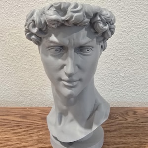 May include: A gray bust of a man with detailed curly hair and a stern expression. The sculpture is set on a wooden surface against a white wall. The bust is a classical art piece.