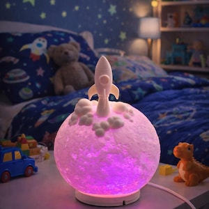 May include: A decorative night light shaped like a moon with a rocket ship on top. The moon lamp emits a soft pink and purple glow. The lamp is on a white base and is designed for a child's bedroom.