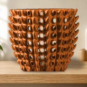 May include: A metallic copper-colored planter featuring a repeating geometric pattern of vertical ridges and circular cutouts. The cylindrical vessel has a textured, spiked exterior surface with a consistent finish throughout the entire structure.