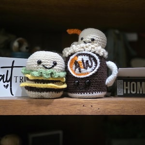 Puede incluir: Juguetes amigurumi de ganchillo: hamburguesa y root beer float. La hamburguesa tiene un pan blanco, lechuga verde, queso amarillo y una hamburguesa marrón. El root beer float tiene una taza marrón con espuma blanca y el logo de A&W.