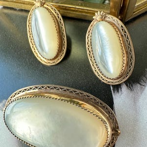 Könnte beinhalten: Vintage Schmuckset mit einer Brosche und passenden Ohrringen. Die ovalen Stücke haben eine perlmuttartige Mitte, die von einem goldfarbenen Filigranmuster umrahmt ist. Die Ohrringe haben ein kleines Kronendetail oben.