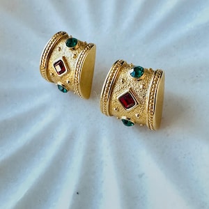 Puede incluir: Un par de pendientes dorados. Cada pendiente presenta un diseño cilíndrico con una superficie texturizada, adornado con una gema cuadrada roja central y gemas redondas verdes. Los pendientes se presentan sobre una superficie blanca.