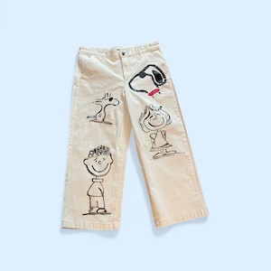 May include: Cream-colored pants with black and red cartoon character illustrations. The wide-leg pants feature a button and zipper closure. The illustrations include a dog, a bird, and a boy.