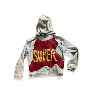 May include: A light-wash denim jacket with a gray hood. The back of the jacket features a red painted design with the word "SUPER" in gold lettering. The jacket has long sleeves and a classic denim design.