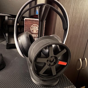 Könnte beinhalten: Ein schwarz-weißes Gaming-Headset ruht auf einem schwarzen Ständer mit einem radförmigen Design. Der Ständer hat einen roten Akzent. Der Hintergrund umfasst einen Computermonitor und einen Holzschrank.
